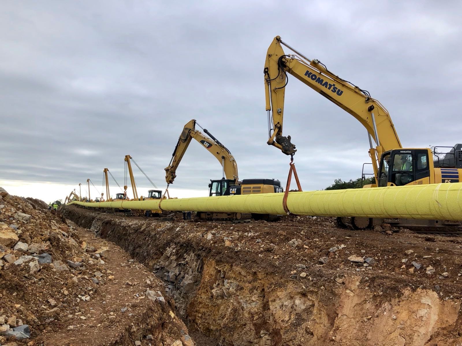 A line of sidebooms and excavators working an open rock trench.