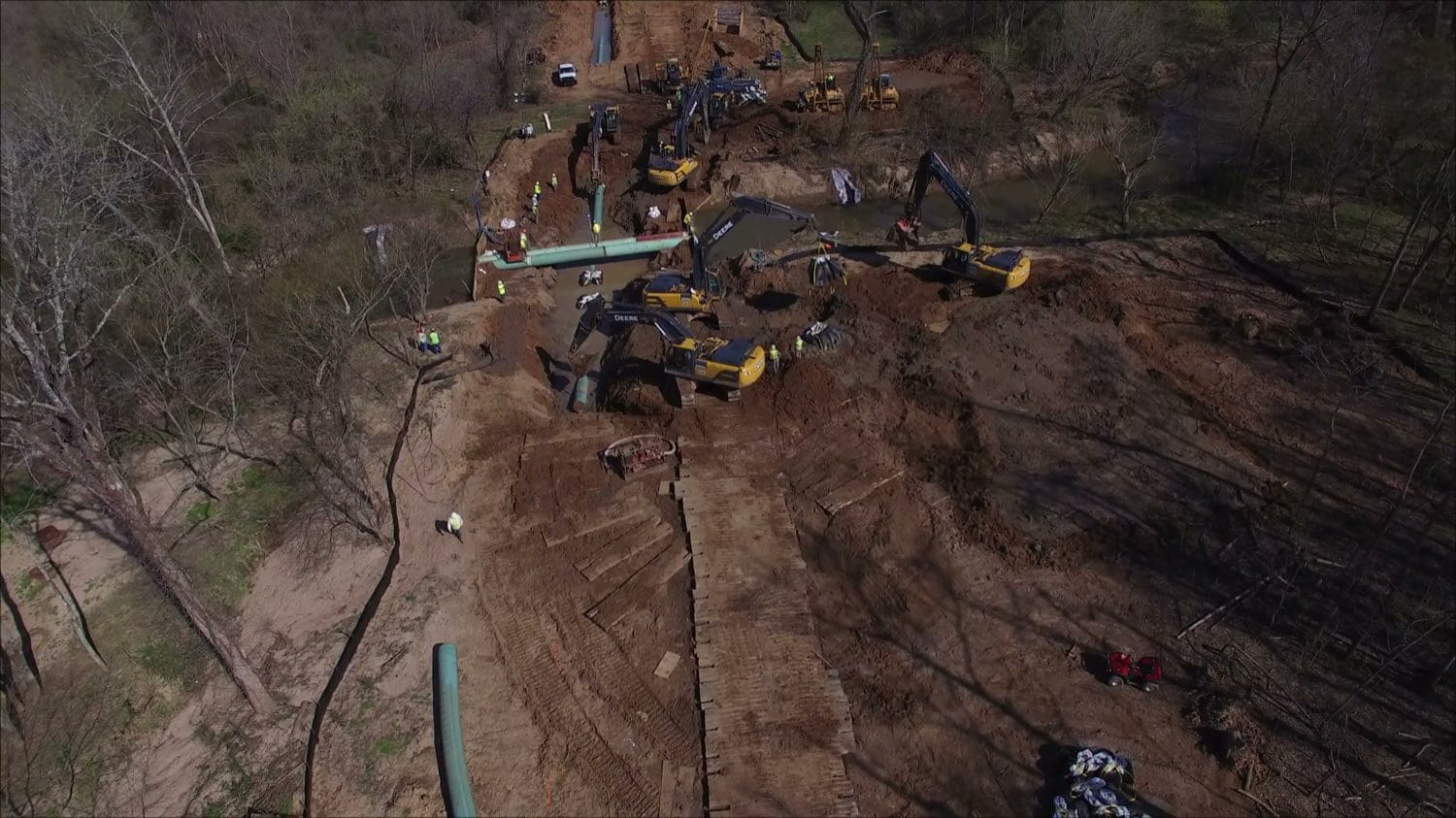 A creek crossing worked by a line of excavators, with coated pipe staged alongside.